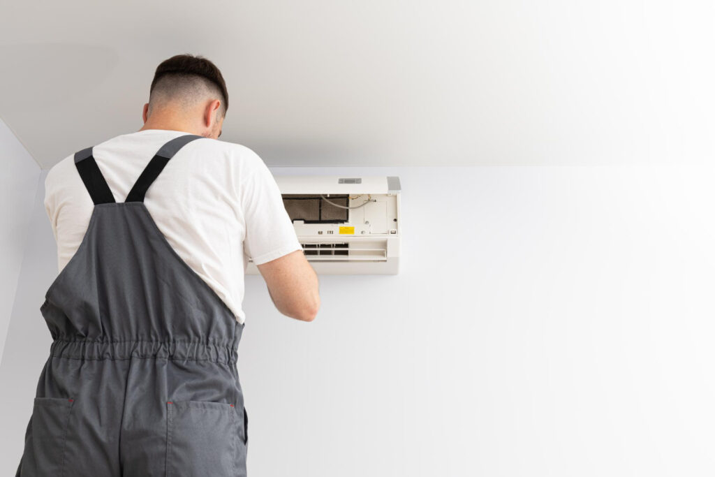 A technician in gray overalls, seen from behind, installs or repairs a wall-mounted air conditioner.