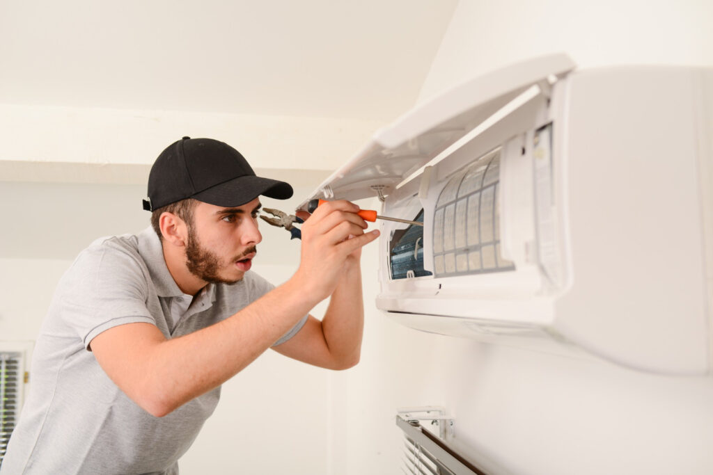 A technician in a cap and gray polo shirt intently repairs an air conditioner with a screwdriver.
