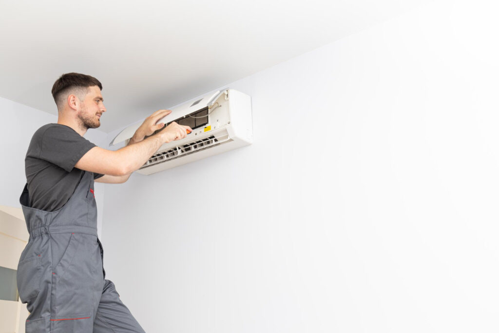 A man in overalls repairs an air conditioner, focused on the unit with tools in hand.