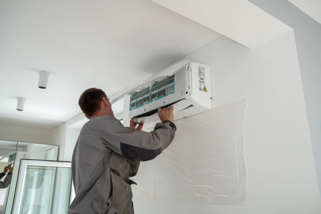 A technician in a gray uniform installs a wall-mounted air conditioner in a room with white walls and ceiling lights.